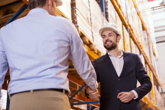 Two businessmen in hard hats standing in warehouse, smiling and shaking hands