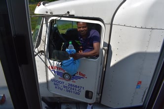 A smiling truck driver looks out the window of the cab while holding his swag bag, snacks, and drink